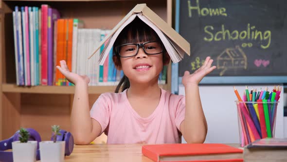 Cute little girl holding a book over her head like a roof, smiling and looking at the camera.