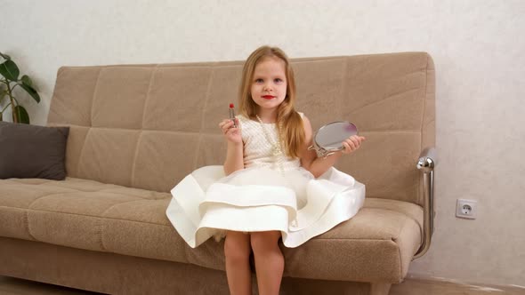 A Young Blonde Little Girl Holds a Mirror and Lipstick in Her Hands