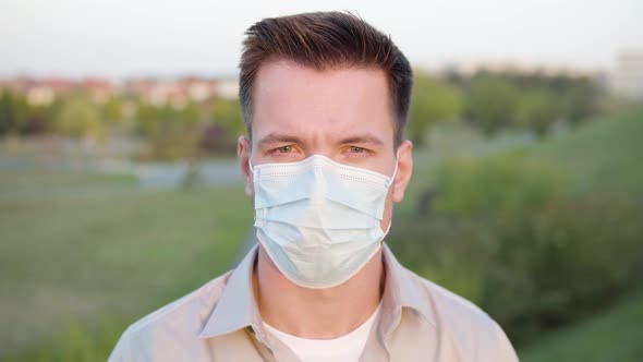 A Young Caucasian Man in a Face Mask Looks at the Camera on a Town's Outskirts  Closeup