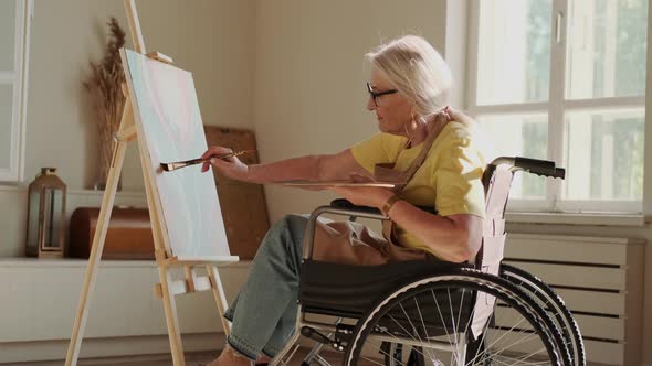 Elderly Disabled Woman Paints a Picture While Sitting in a Wheelchair in a Workshop