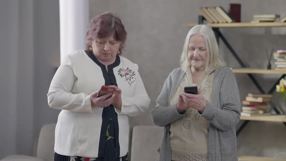 Front View Portrait of Two Absorbed Senior Caucasian Women Using Smartphones Standing in Living Room