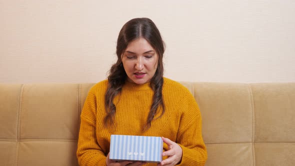 Beautiful Young Woman Sitting on the Sofa Opens a Gift and is Very Disappointed