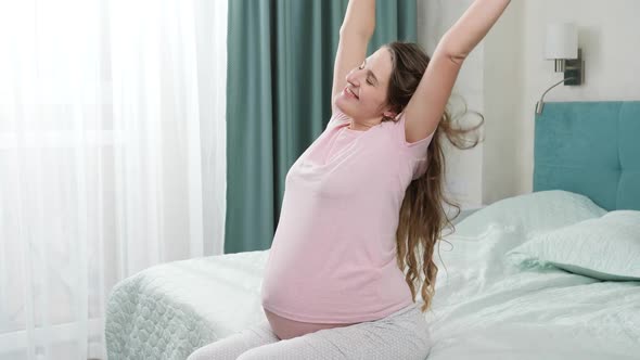 Portrait of Smiling Happy Pregnant Woman in Pajamas Stretching on Bed at Morning After Waking Up