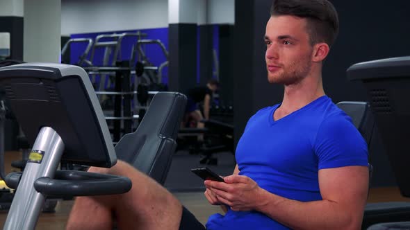 A Young Fit Man Trains on a Recumbent Bike in a Gym and Works on a Smartphone - Closeup