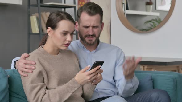Portrait of Couple Reacting to Loss on Smartphone Sitting on Sofa