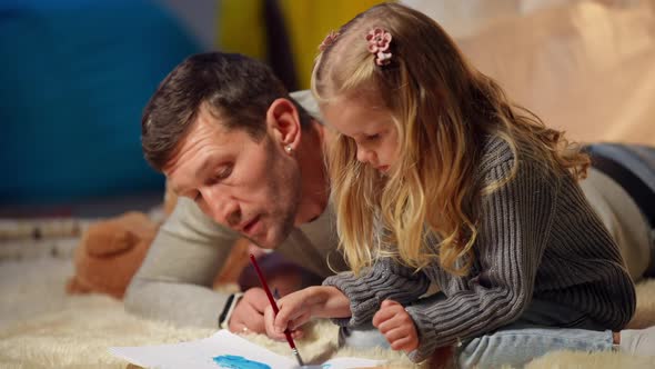 Side View Focused Talented Little Girl Painting with Blurred Curios Man Lying at Background at Tent