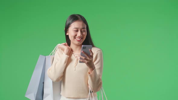 Asian Shopping Woman Holding Shopping Bags Use Mobile Phone And Smile While Walking On Green Screen