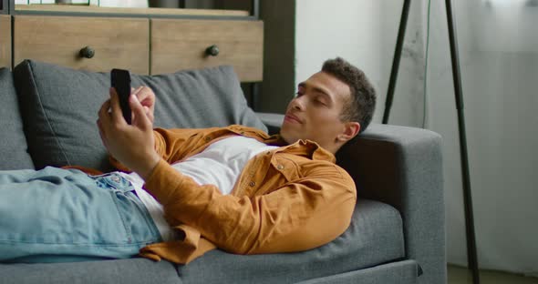 Handsome Hispanic Man Is Lying on Sofa, Using His Smartphone