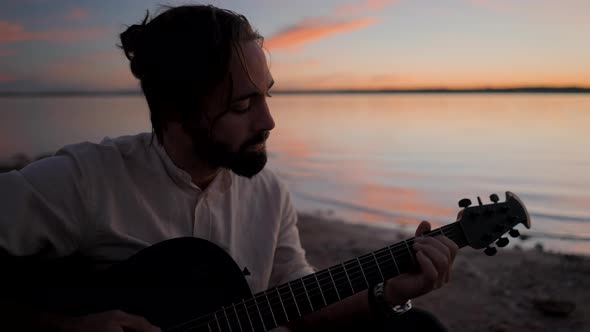 Thoughtful Bearded Hipster Man Playing an Acoustic Guitar By the Lake Against a Sunset Sky