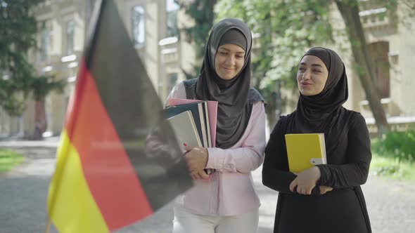 Two Female Muslim Immigrants Standing with Book on University Yard and Talking As Blurred German