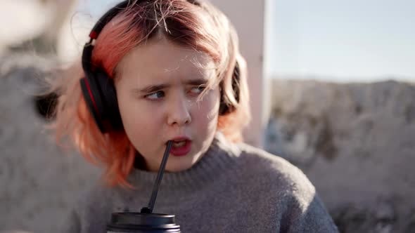 Cute Little School Girl is Listening to Music By Headphones and Drinking Cocktail By Straw on Street