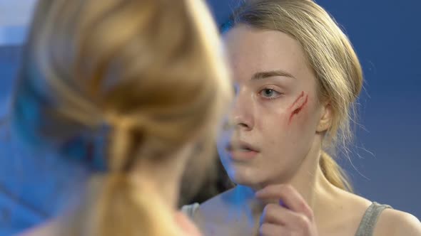 Sad Teenager Girl Looking at Wounded Face in Mirror, Father Tyrant on Background