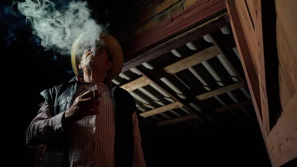 An elderly farmer in a straw hat smokes a cigar outside the ranch. The man smokes a cigar