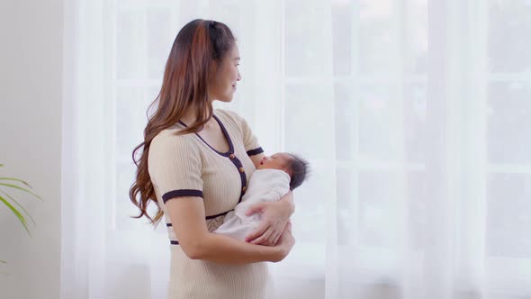 Asian woman holding newborn baby in her arms standing in front of windows