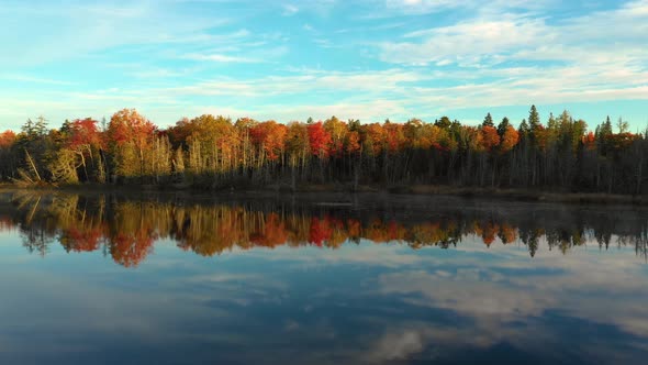 Aerial footage sliding left across glassy pond with forested shore in fall colors