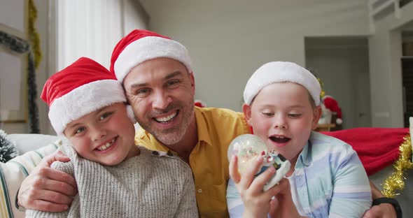 Portrait of caucasian father and two sons wearing santa hats waving and smiling sitting on the couch