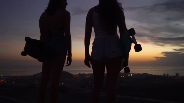 Two Girls Standing on the Edge of the Road with Skateboards in Their Hands Watching the Sunset