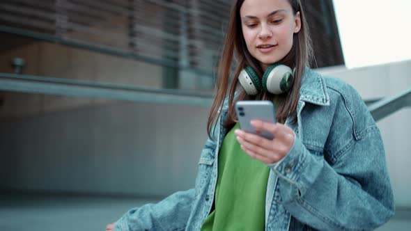 Pensive blonde woman in headphones wearing jeans jacket typing by phone and walking