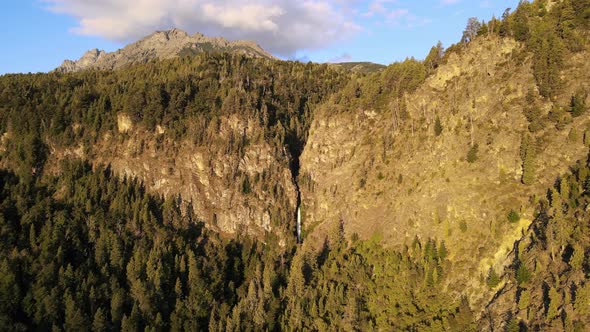 Aerial orbit of Corbata Blanca waterfall hiding between steep pine tree forest mountains at golden h