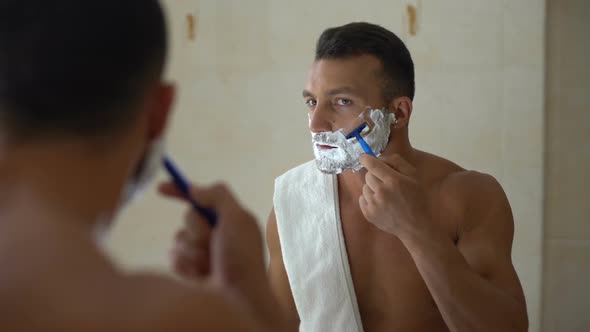 Male With Foam on Face Using Manual Razor to Trim and Shave Beard in Bathroom