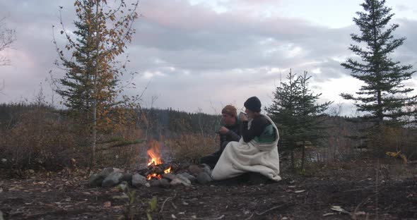 Couple Sitting by Campfire on Camping Trip in WIlderness of Alaska and Drinking From Cups in Twiligh