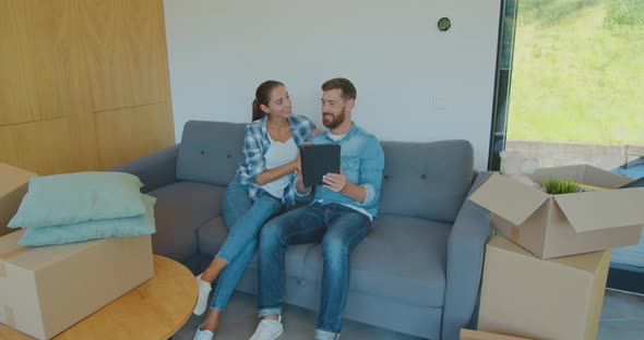 Happy Couple Renters Owners Tenants Sit on Sofa Use Digital Tablet on Moving Day in New House, Man