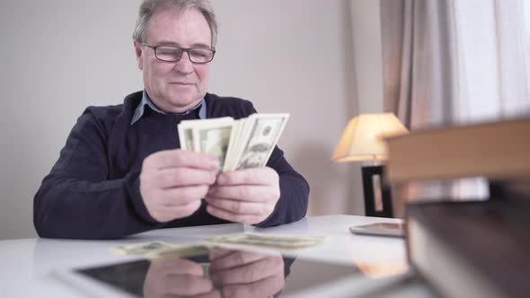 Portrait of Rich Old Caucasian Man Counting Cash Indoors. Smiling Senior Male Retiree in Eyeglasses