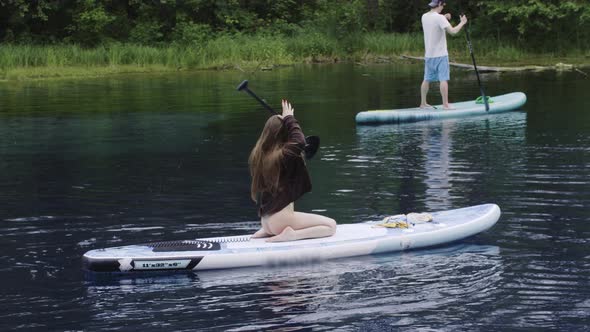 Young Brunette Woman Sitting on Her Knees on the Boards in the River and Twists the Paddle Over Her