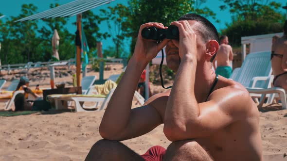 Man on the Beach in Swimming Trunks is Watching Someone Through Binoculars