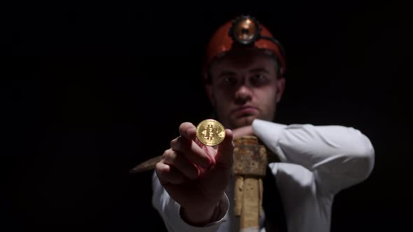 Caucasian Young Man in Hard Hat is Leaning on a Pickaxe and Holding Shiny Bitcoin Coin at Arm's