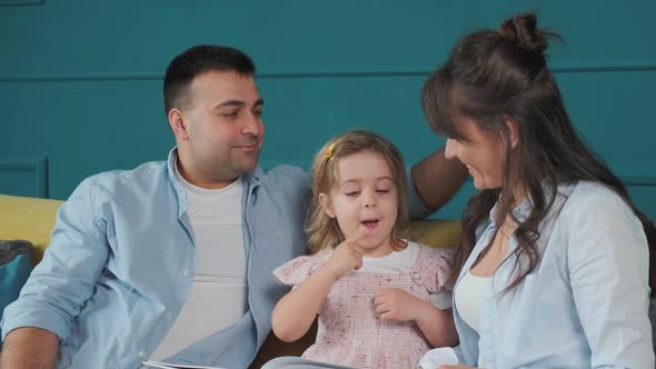 Young Family Reads a Book to Their Little Daughter While Sitting on the Couch