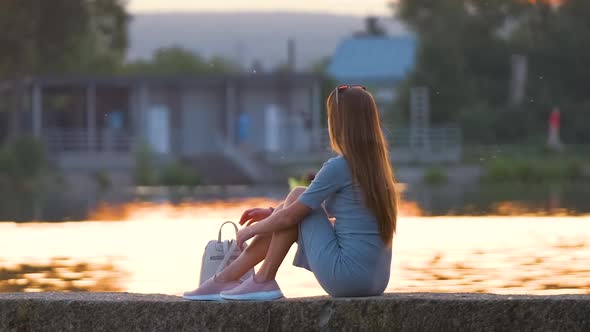 Back View of Lonely Woman Sitting on Lake Shore on Warm Evening