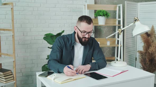 Bearded Guy Learn Online Courses Using a Tablet and Write Notice in His Notebook