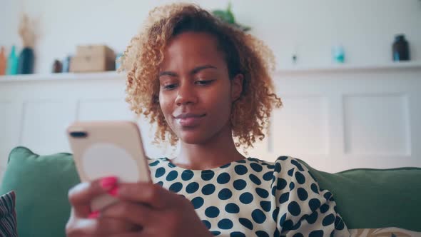 Young Pretty African American Woman with Mobile Phone in Hands Sits on Sofa