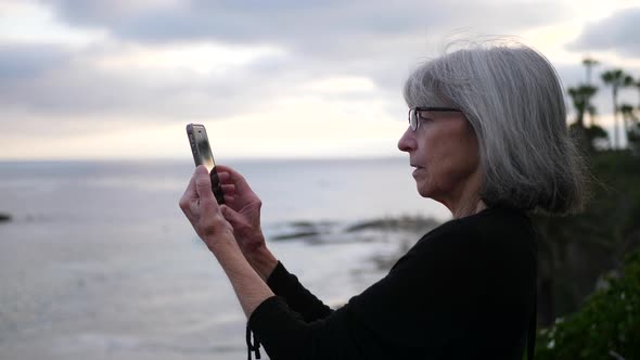 An old aging woman on vacation using her phone to take a picture at the beach over the ocean at suns