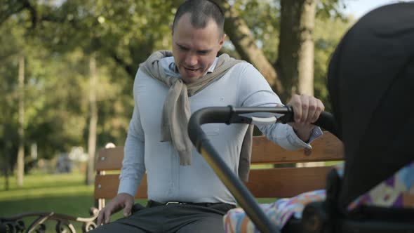 Portrait of Exhausted Young Caucasian Man Yawning and Rocking Baby Stroller Outdoors. Tired Father