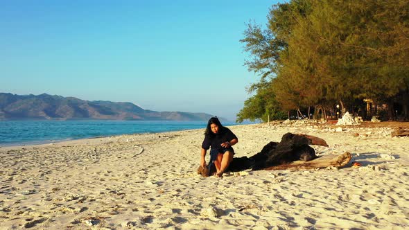 Girl sitting on a trunk playing with white sand of exotic beach washed by blue sea surrounding tropi