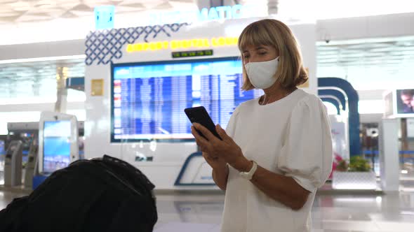 Middle Aged Woman Wearing Face Mask Checking Her Phone Standing By the Departure Board at the