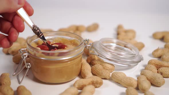 A hand takes a dessert with a spoon, juicy peanut paste with nuts from a glass jar on a white table