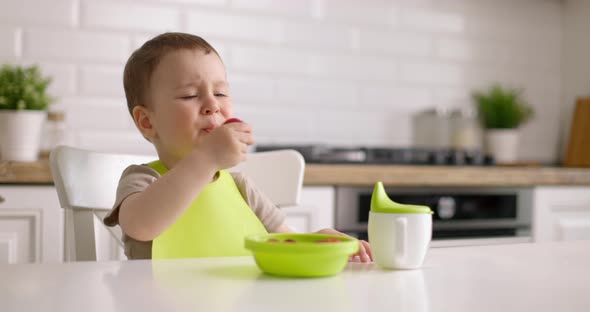 Cute Baby Boy Sits at Table in the Kitchen and Eats Berries with His Hand