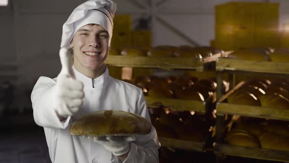 Young Baker Holds a Fresh Loaf of Bread in a Hand and Shows a Thumb at Camera