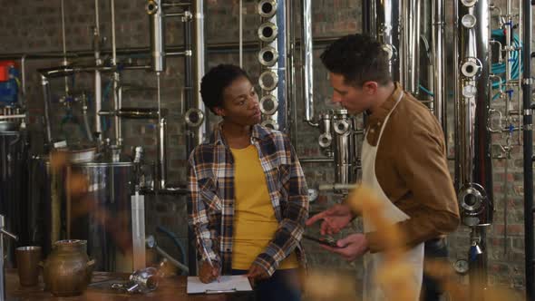 Diverse male and female colleague at gin distillery checking equipment, using tablet and discussing