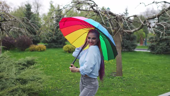 A Cute Girl with Multicolored Braids and Bright Makeup in a Bluish Shirt Posing with a Rainbow