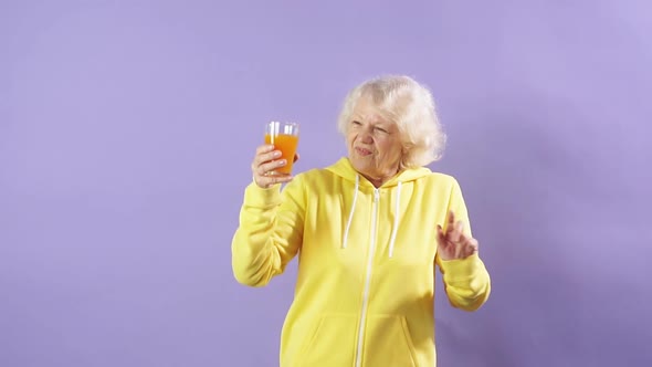 Joyful Old Woman in a Yellow Sports Sweatshirt Holds a Glass of Fresh Carrot Juice in Her Hand