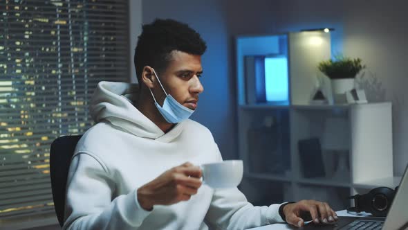 Medium Shot of Handsome Multiracial Man Drinking a Cup of Coffee and Working on Computer at Night
