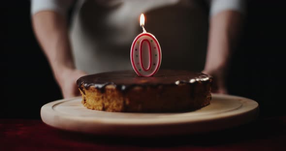 Young Woman Wearing Kitchen Apron Puts Homemade Chocolate Cake or Pie with Number 0 Burning Candle