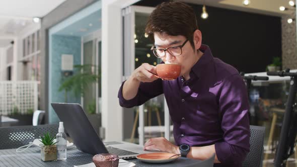 Asian man with laptop drinking coffee while sitting at a cafe