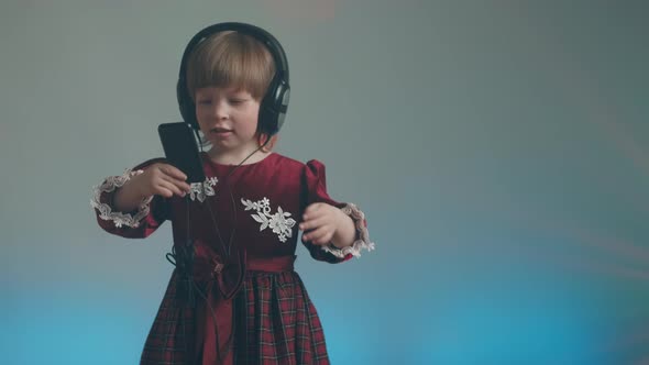 Little Girl in Big Headphones and Vintage Dress is Listening to Music on Phone