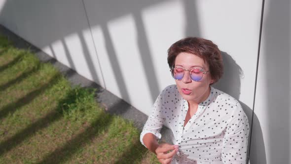 Woman with Colorful Sunglasses Sits on Lawn in Urban Park and Enjoys of Bright Sunlight