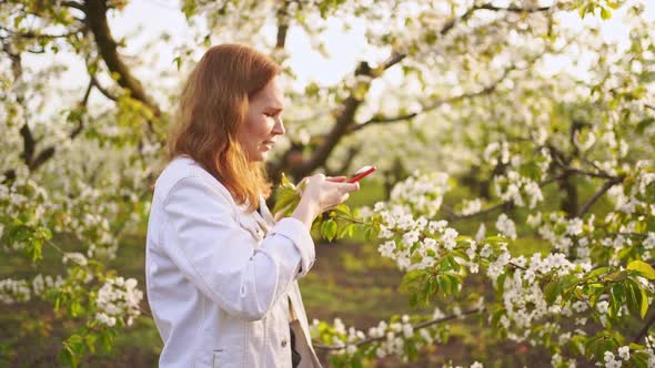 A Woman in a White Jacket Shoots a Video on Her Phone in a Flowering Garden